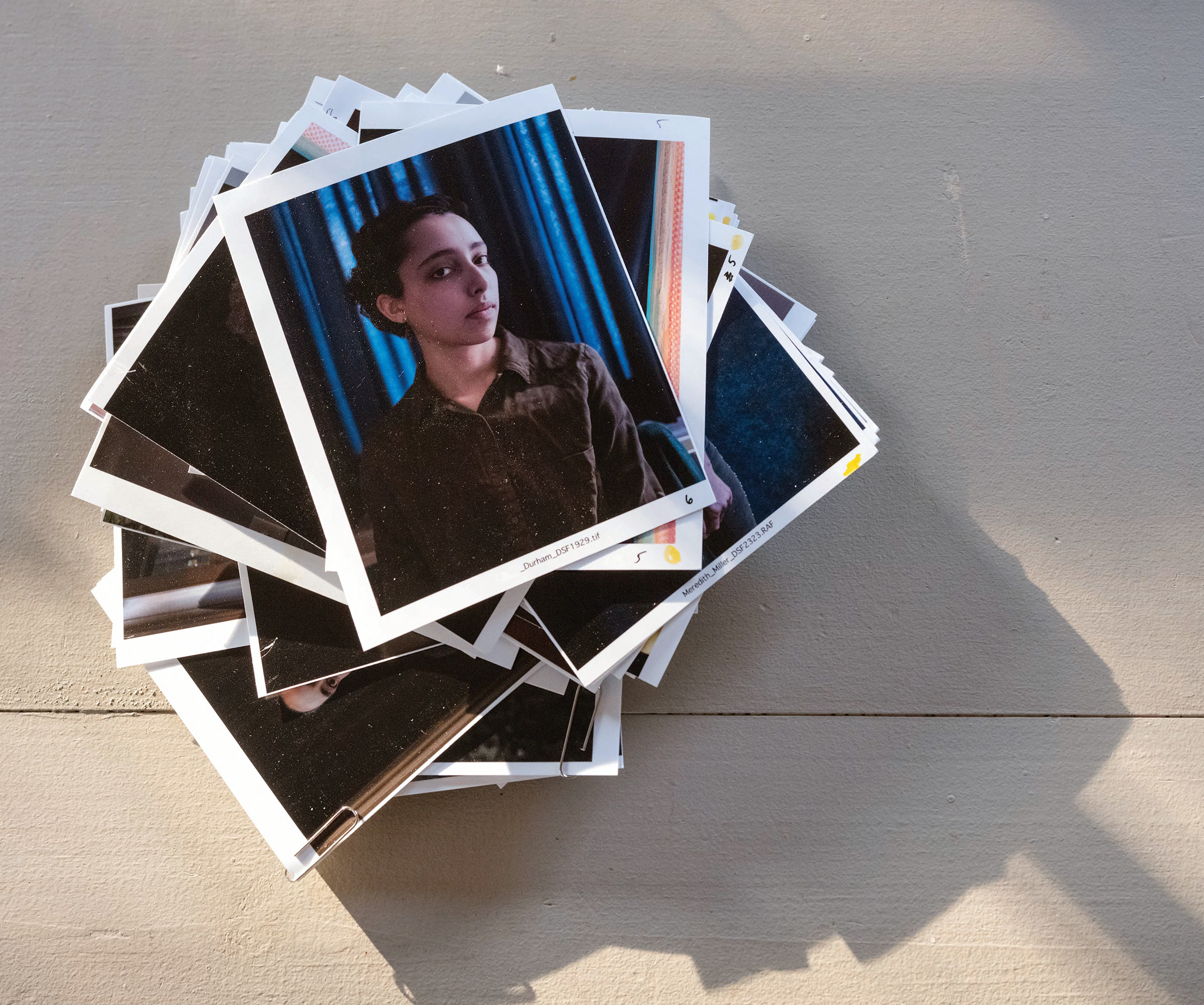 An overhead image of a stack of small contact sheets in disarray featuring individual portraits, set on a table with bands of  light and shadow.